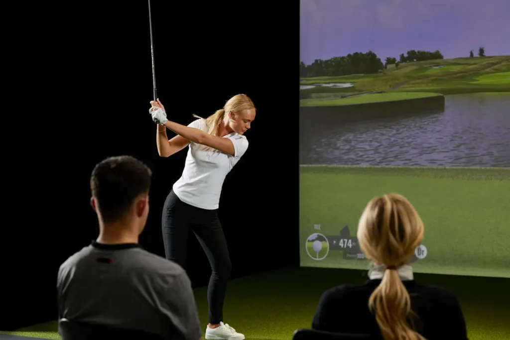 Golfer using an indoor golf simulator with spectators watching in a clubhouse coaching and training environment.